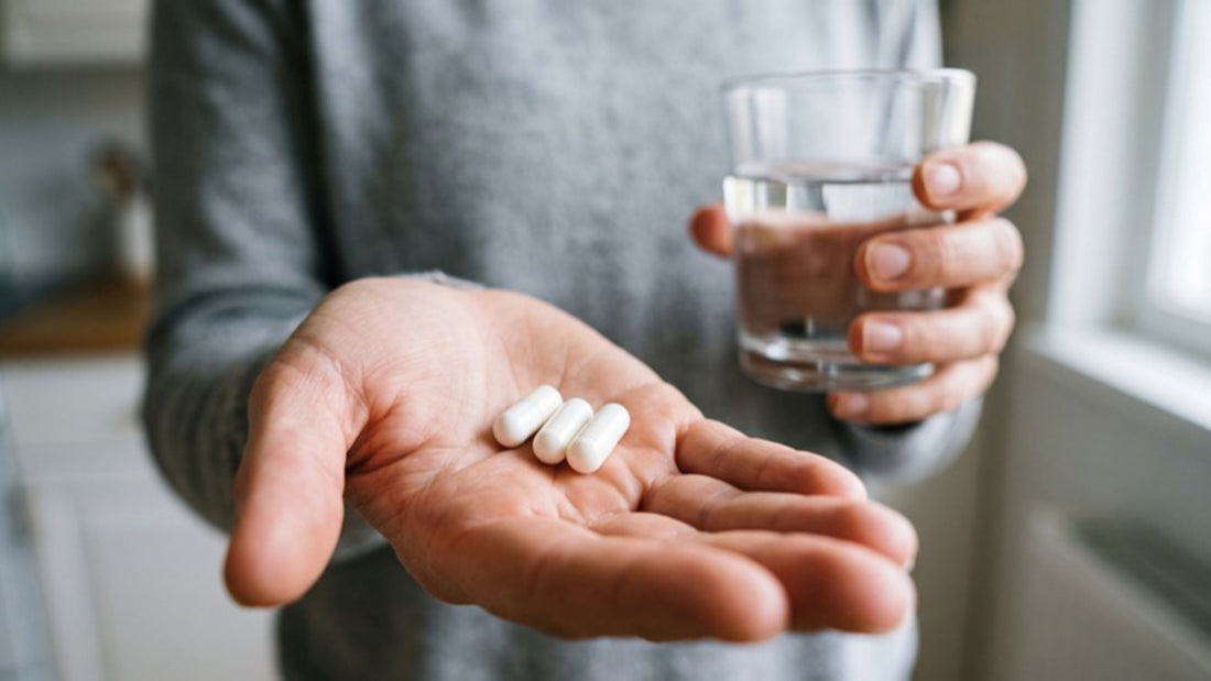 Offene Hand mit drei weissen Kapseln, in der anderen Hand ein Glas Wasser